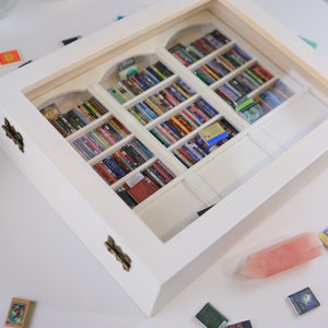 Może przedstawiać: Białe drewniane pudełko ze szklanym frontem, prezentujące miniaturową biblioteczkę wypełnioną kolorowymi książkami. Pudełko ma dwa mosiężne zawiasy, a na stole poniżej znajduje się r&oacute;żowy kryształ.