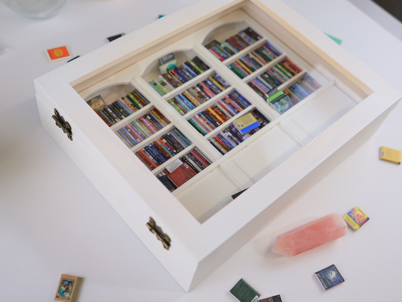 Może przedstawiać: Białe drewniane pudełko ze szklanym frontem, prezentujące miniaturową biblioteczkę wypełnioną kolorowymi książkami. Pudełko ma dwa mosiężne zawiasy, a na stole poniżej znajduje się r&oacute;żowy kryształ.