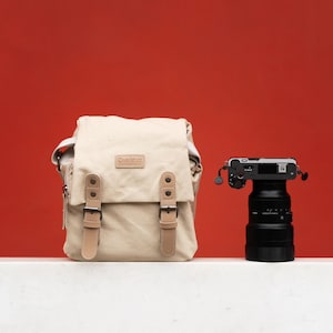May include: A beige canvas camera bag with leather details and a black camera with a lens. The bag has a flap closure with two buckle straps. The camera is on a black lens, set against a red background.