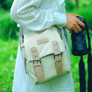 Peut inclure: Un sac d'appareil photo en toile beige avec une fermeture à rabat et deux sangles en cuir avec boucles. Le sac a une bandoulière et un petit patch en cuir avec le mot "cwatsun".