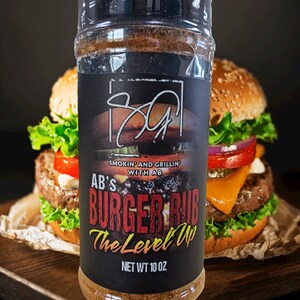 Puede incluir: Un frasco transparente de AB's Burger Rub con una etiqueta negra que presenta el texto "Smokin' and Grillin' with AB" y "Burger Rub The Level Up". El frasco contiene una mezcla de especias marrones. Peso neto 283 gramos.
