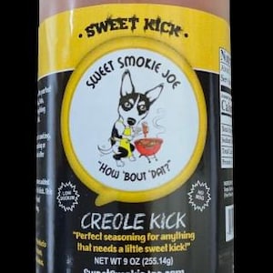 May include: A cylindrical container of Creole Kick seasoning. The label features a cartoon dog chef with a barbecue, the text "Sweet Smokie Joe", and the phrase "How 'bout dat?" The net weight is 9 oz (255.14g).