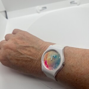 May include: White wristwatch with a colorful watercolor design on the face. The watch has a white band and a round face. The watch is on a wrist.
