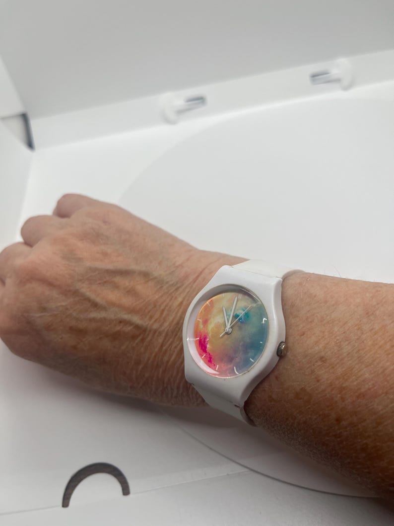 May include: White wristwatch with a colorful watercolor design on the face. The watch has a white band and a round face. The watch is on a wrist.