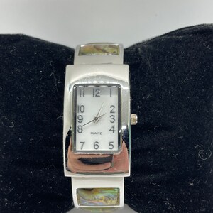 May include: A silver rectangular watch with a white face and black hands. The watch has a mother-of-pearl band with a silver buckle. The watch face says "QUARTZ" at the bottom.