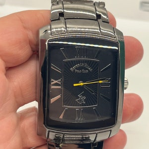 May include: A black and silver men's watch with a rectangular face and Roman numeral hour markers. The watch has a black band with a silver buckle. The watch face has a black background with a silver logo and the words "Forever Yours Polo Club".