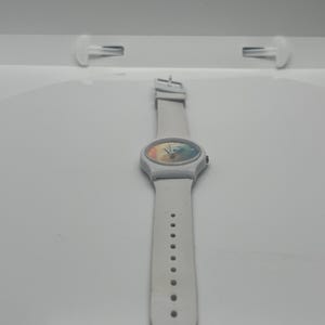 May include: A white wristwatch with a colorful face and a white band. The watch has a round face with a rainbow-like design. The band has multiple holes for adjustment.