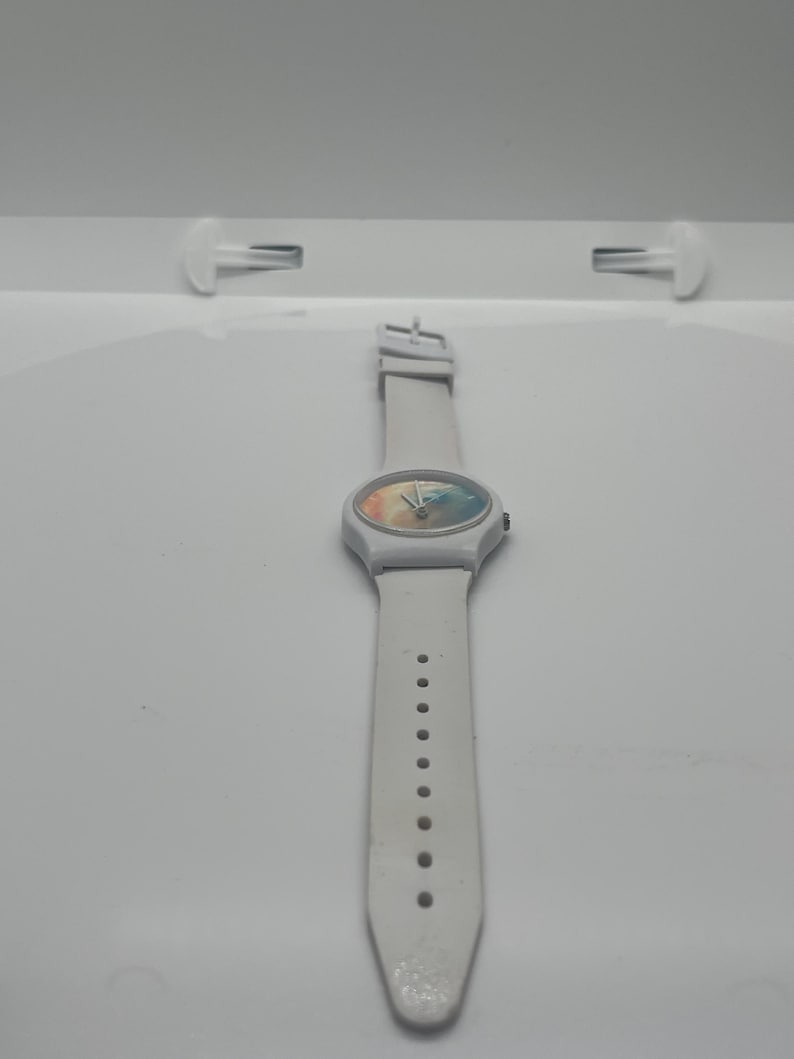 May include: A white wristwatch with a colorful face and a white band. The watch has a round face with a rainbow-like design. The band has multiple holes for adjustment.
