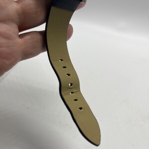May include: A black leather watch strap with a brown leather lining and stitching. The strap has multiple holes for adjusting the fit.