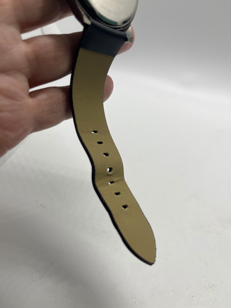 May include: A black leather watch strap with a brown leather lining and stitching. The strap has multiple holes for adjusting the fit.