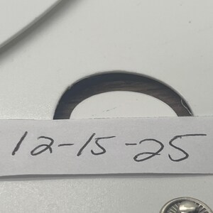 May include: Close-up of a white card with the handwritten numbers "12-15-25" in black ink. A silver snap is visible at the bottom right. The card has a circular cutout at the top.