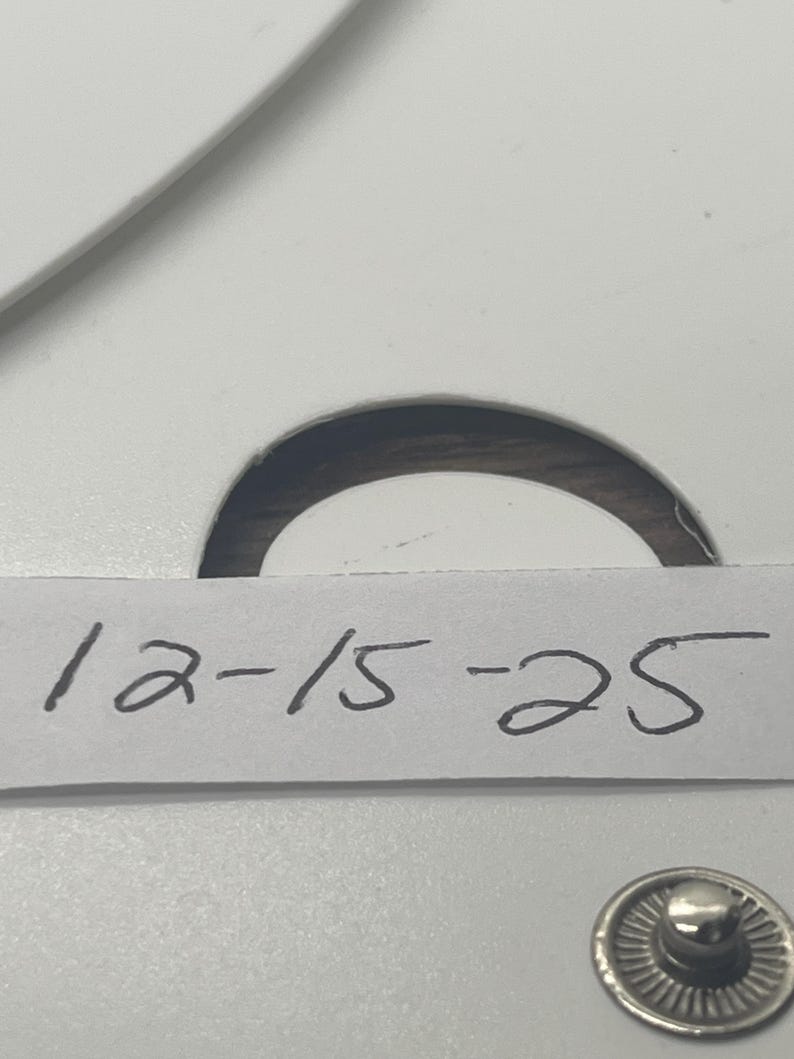 May include: Close-up of a white card with the handwritten numbers "12-15-25" in black ink. A silver snap is visible at the bottom right. The card has a circular cutout at the top.