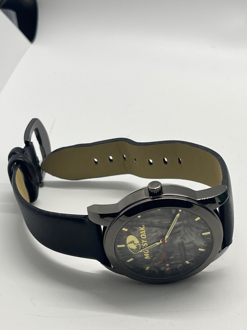 May include: Black leather wristwatch with a black face and a camouflage pattern. The watch face has a gold "MOssy OAK" logo.