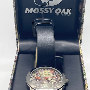 May include: A black wristwatch with a black leather strap and a camouflage face. The watch is in a box with the Mossy Oak logo and the words "Brand Camo".