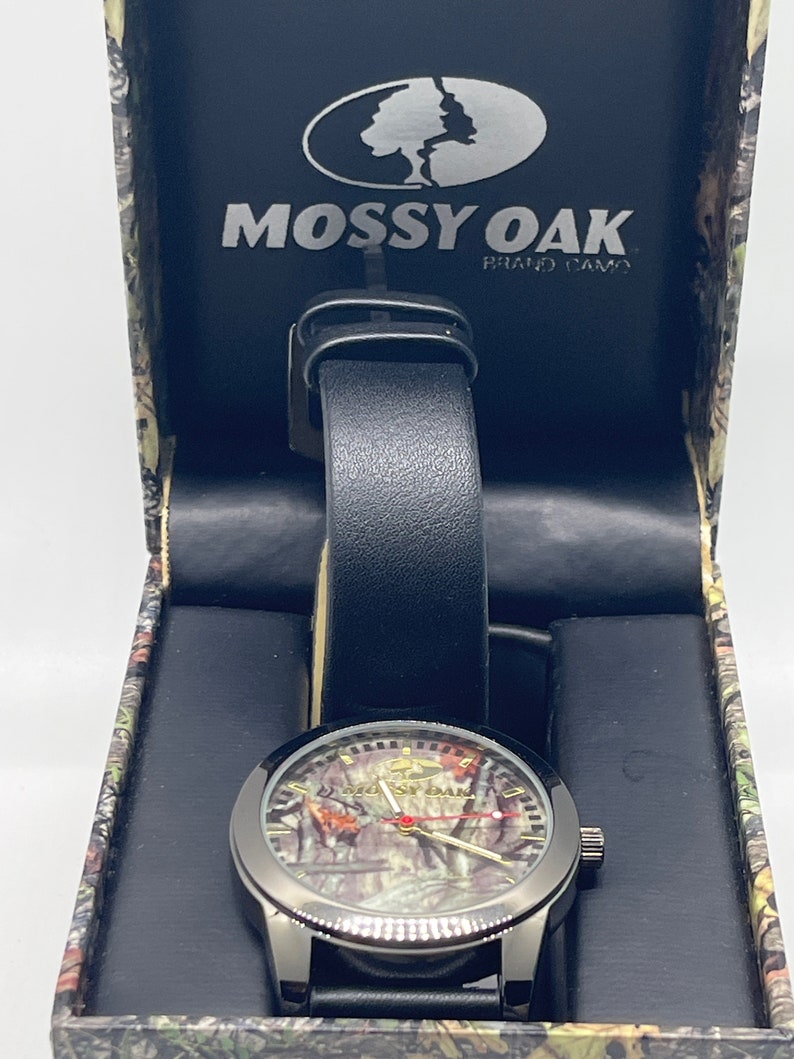 May include: A black wristwatch with a black leather strap and a camouflage face. The watch is in a box with the Mossy Oak logo and the words "Brand Camo".