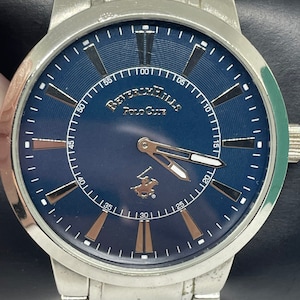May include: A silver wristwatch with a blue face and silver hands. The face has a white ring with black markings for the minutes and a gold ring with Roman numerals for the hours. The watch face also has the text "Beverly Hills Polo Club" in gold lettering.
