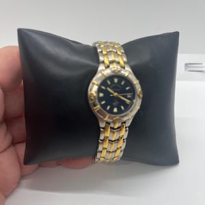 May include: A silver and gold-tone wristwatch with a dark blue face. The watch has a gold-tone bezel and a two-tone metal band. The watch is displayed on a black pillow.