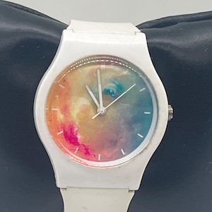 May include: White wristwatch with a colorful, abstract design on the watch face. The watch has a white band and a silver crown. The watch face features a blend of pink, orange, blue, and green hues, creating a vibrant, artistic look.