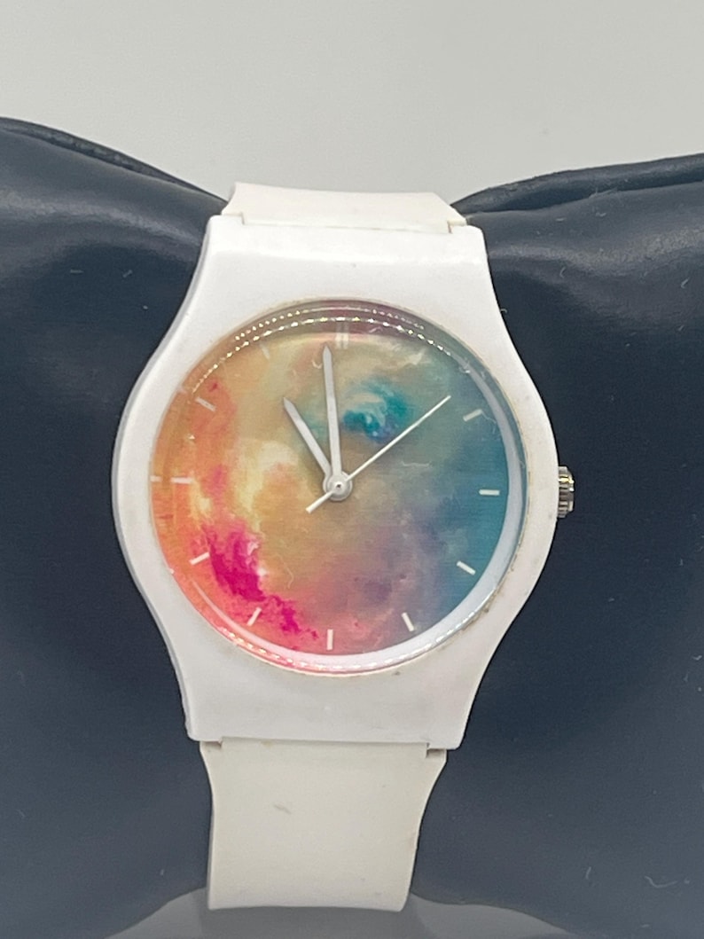 May include: White wristwatch with a colorful, abstract design on the watch face. The watch has a white band and a silver crown. The watch face features a blend of pink, orange, blue, and green hues, creating a vibrant, artistic look.