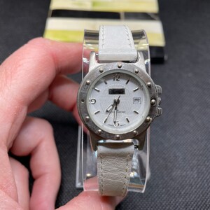 May include: A silver wristwatch with a white face and a white leather band. The watch has a date window and a silver bezel with small dots around the edge.