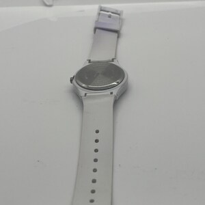 May include: A white wristwatch with a white strap. The watch face is round with a silver-colored back. The strap has a buckle and several holes for adjustment. The watch is set against a white background.