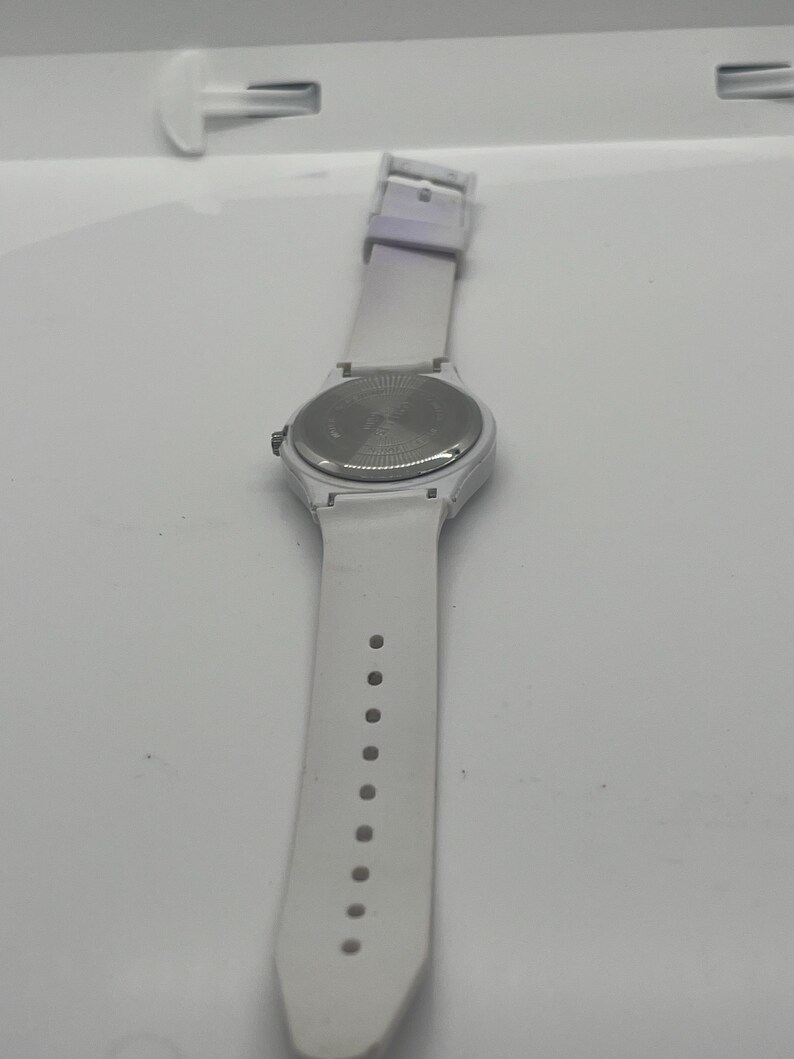 May include: A white wristwatch with a white strap. The watch face is round with a silver-colored back. The strap has a buckle and several holes for adjustment. The watch is set against a white background.