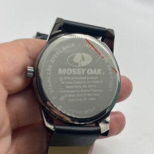 May include: A black leather strap watch with a silver face. The watch face features the Mossy Oak logo and text that reads "1ATM Water Resistant, Mossy Oak, Official licensed product of Haas Outdoors, Inc. &copy;2018 West Point, MS 39773, Distributed by Xtreme Time Inc. 20 West 33rd St. 6th Floor, New York, NY 10001, Japan Movt."
