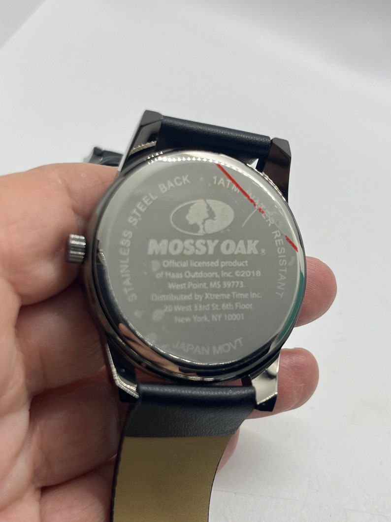 May include: A black leather strap watch with a silver face. The watch face features the Mossy Oak logo and text that reads "1ATM Water Resistant, Mossy Oak, Official licensed product of Haas Outdoors, Inc. &copy;2018 West Point, MS 39773, Distributed by Xtreme Time Inc. 20 West 33rd St. 6th Floor, New York, NY 10001, Japan Movt."