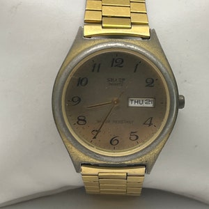 May include: A gold-toned wristwatch with a round face and a gold metal band. The watch face has black Roman numeral hour markers, black hands, and a date window that displays "THU 21". The watch face also has the text "SHARP QUARTZ" and "WATER RESISTANT".