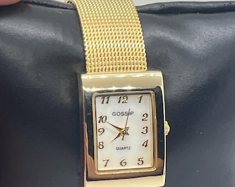 May include: Gold-toned rectangular watch with a mesh band. The watch face is white with black numerals and hands, and the word "GOSSIP" is printed above the hands. The watch is a quartz movement.