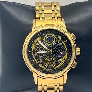 May include: A gold-tone analog wristwatch with a metal link band. The black dial features a skeletonized design revealing gold mechanical gears, Roman numeral hour markers, white hands, a moon phase sub-dial, and a small date display window.