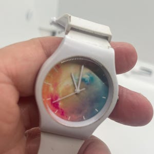May include: White wristwatch with a colorful, abstract design on the watch face. The watch has a white band and a round face with a mix of pink, yellow, blue, and orange hues. The hands are silver.