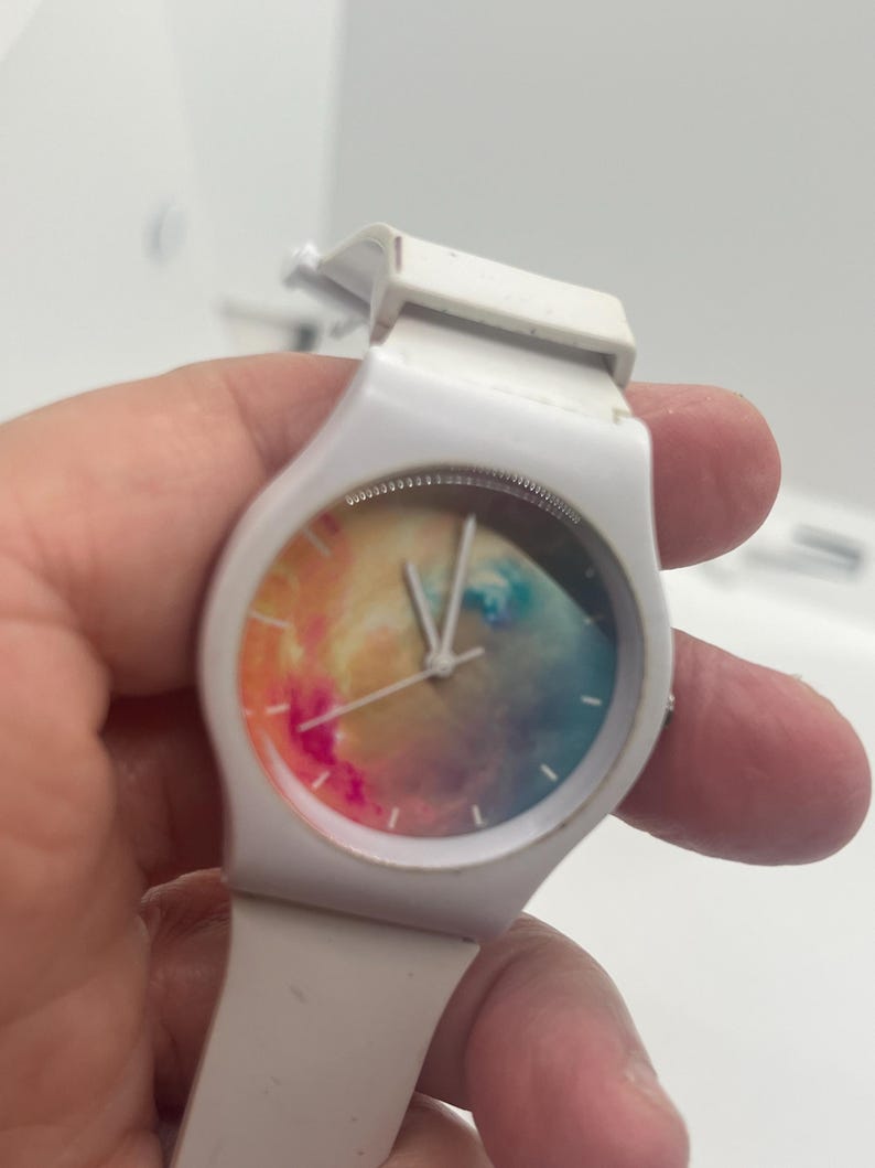 May include: White wristwatch with a colorful, abstract design on the watch face. The watch has a white band and a round face with a mix of pink, yellow, blue, and orange hues. The hands are silver.