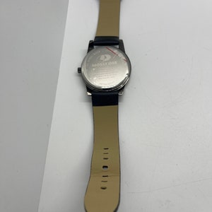 May include: A black leather wristwatch with a silver face and a stainless steel back. The watch face has the words "Mossy Oak" printed on it. The watch has a black leather strap with a buckle closure.