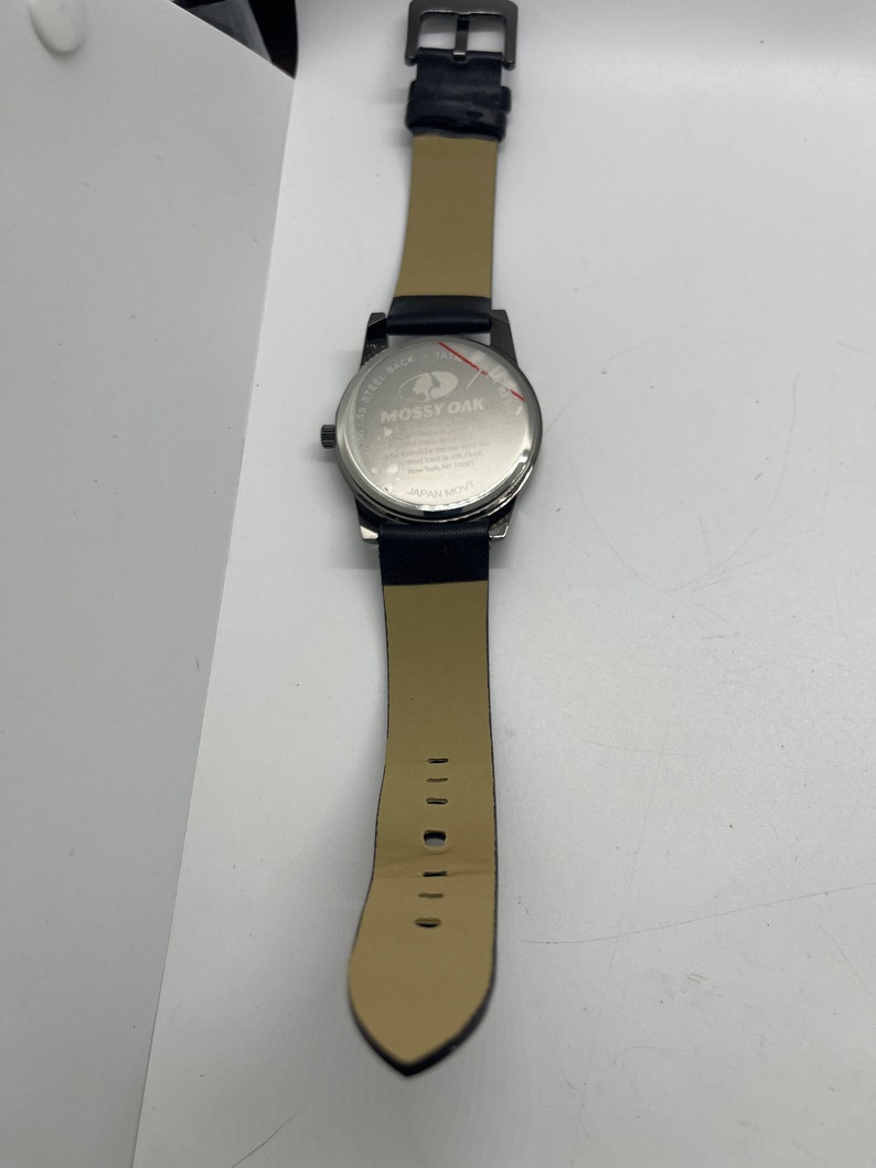 May include: A black leather wristwatch with a silver face and a stainless steel back. The watch face has the words "Mossy Oak" printed on it. The watch has a black leather strap with a buckle closure.