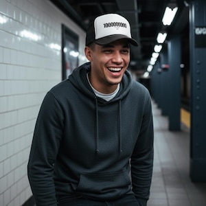 Puede incluir: Gorra de camionero en blanco y negro con el texto "10000000000" en negro. La gorra tiene una visera negra y un panel frontal blanco. La persona de la imagen lleva una sudadera con capucha gris oscuro.