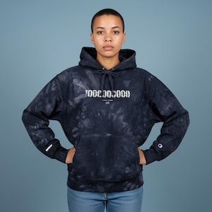 Może przedstawiać: Granatowa bluza z kapturem typu tie-dye z białym napisem "1000000000". Bluza posiada kaptur ze sznurkiem i logo Champion na lewym rękawie. Modelka ma na sobie niebieskie dżinsy.