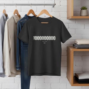 May include: Dark grey t-shirt displaying the number "10000000000" in white, accompanied by the text "Street Code 1979 2020". The t-shirt is suspended on a wooden hanger, with other garments visible in the background.