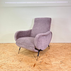 Könnte beinhalten: Ein plüschiger, mauvefarbener Sessel mit einer geschwungenen Rückenlehne und Armlehnen. Der Sessel hat schwarze Metallbeine mit goldenen Akzenten. Der Sessel steht auf einem Holzboden.