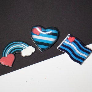 May include: Three blue and white striped pins with red hearts. One pin is a heart shape, one is a rainbow, and one is a flag.
