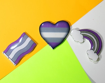May include: Three colorful pins on a multi-colored background. The pins are shaped like a heart, a flag, and a rainbow. The heart is purple, white, and gray. The flag is purple, white, and gray. The rainbow is purple, white, gray, and black.