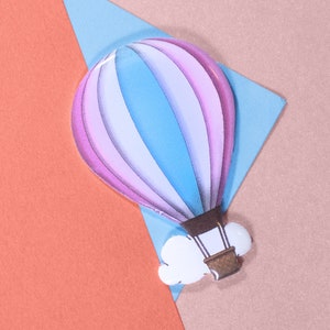 Może przedstawiać: Różowo-niebieski balon na gorące powietrze z białą chmurą i brązowym koszem. Balon znajduje się na różowym, niebieskim i pomarańczowym tle.