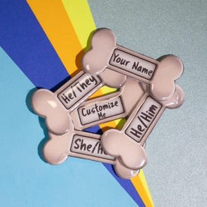 May include: Five bone-shaped tags with different text on them. The tags are beige and have black text. The text on the tags reads "Your Name", "He/They", "Customize Me", "She/He", and "He/Him".