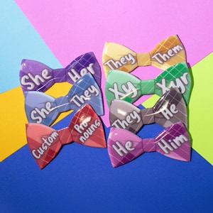 Puede incluir: Un conjunto de ocho pines de corbata de moño con varias opciones de pronombre, incluyendo "She", "Her", "They", "Xy", "Xyr", "He", "Him" y "Custom Pronouns". Los pines están dispuestos en una pila sobre un fondo colorido.