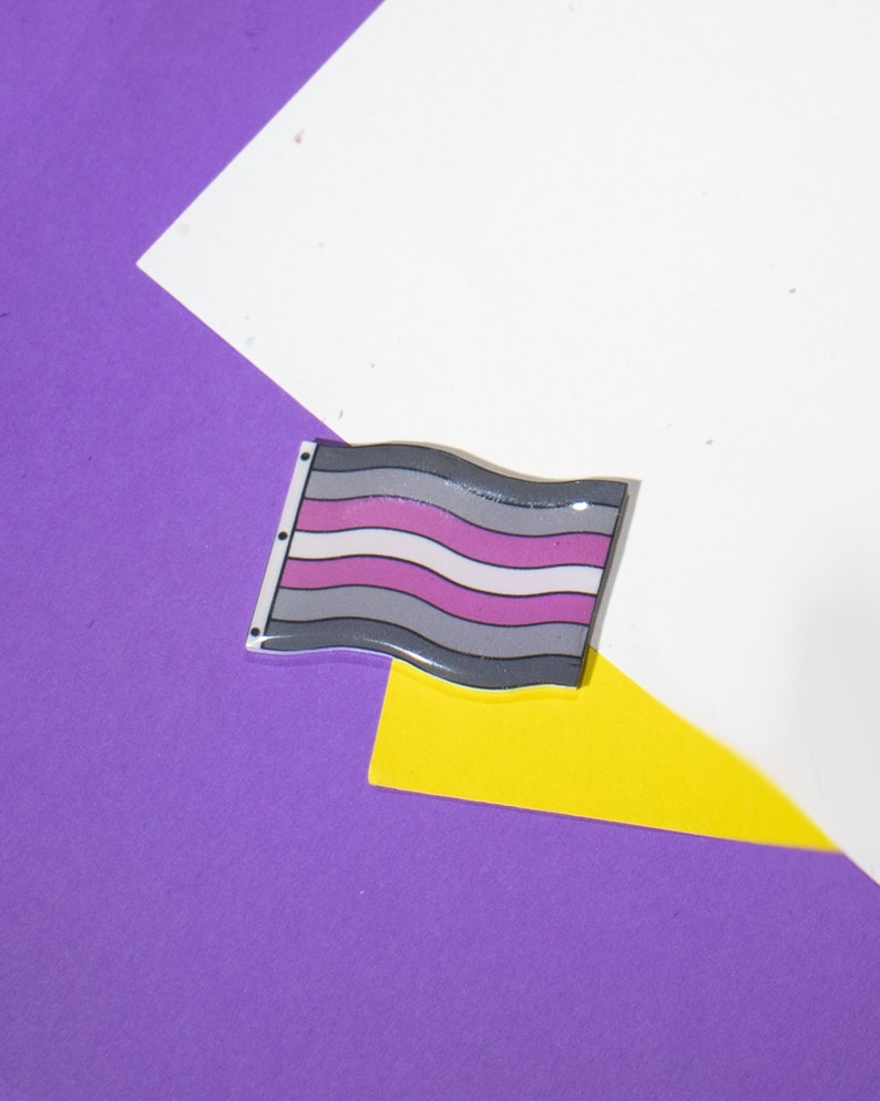 Op de afbeelding: Een grijze, witte en paarse vlag met golvende strepen. De vlag bevindt zich op een paarse en gele achtergrond.