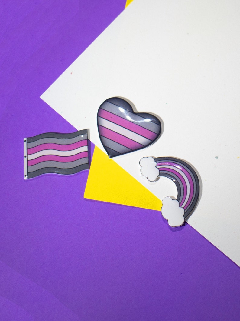 Op de afbeelding: Drie pinnen met een gestreept ontwerp in grijs, wit en roze. De pinnen zijn in de vorm van een hart, een vlag en een regenboog. De pinnen bevinden zich op een paarse, gele en witte achtergrond.