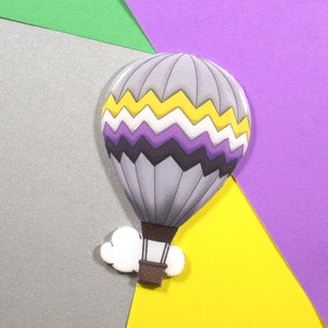 Peut inclure: Un ballon à air chaud gris avec des motifs en zigzag jaunes, blancs, violets et noirs. Le ballon a un panier marron et un nuage blanc attaché. Le fond est composé de formes géométriques grises, vertes, jaunes et violettes.