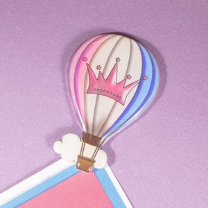 Puede incluir: Un globo aerostático rosa y azul con una corona rosa y estrellas blancas. El globo está unido a una nube blanca y tiene una cesta marrón.