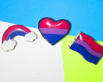 May include: Three enamel pins with a bisexual pride theme. A rainbow, a heart, and a flag.