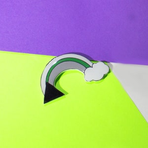 May include: A silver-colored pin shaped like a rainbow with a black triangle at the bottom and a white cloud at the top. The rainbow has green, gray, and white stripes.